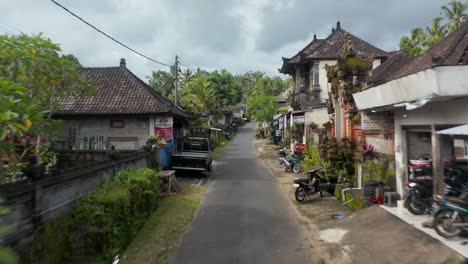 Tiro-Aéreo-De-Carro-De-Bajo-Vuelo-Conduciendo-Por-La-Calle-Tradicional-En-El-Barrio-Rodeado-De-Casas-Residenciales-Y-Templos-En-Bali,-Indonesia