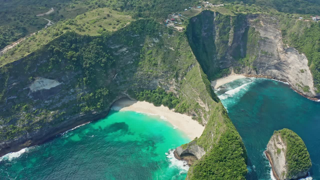 Aerial drone shot in the iconic Kelingking Beach, the 'T-Rex' shaped headland, and the towering limestone cliffs of Nusa Penida. Turquoise Indian Ocean crashes against the rocks and white-sand beach