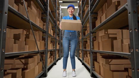 full body of asian female courier in blue uniform showing a carton to camera and smiling while delivering it in warehouse