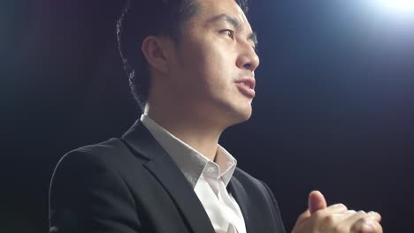 close up side view of asian speaker man in business suit holding his hands together and showing index fingers up while speaking in the black screen studio