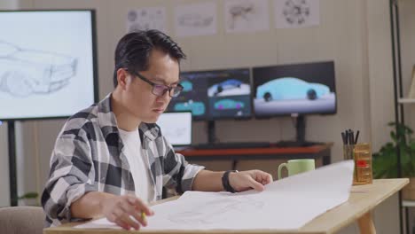 side view of satisfied asian male holding and looking at car design sketch then shaking his head while working on table in the studio with tv and computers display 3d electric car model