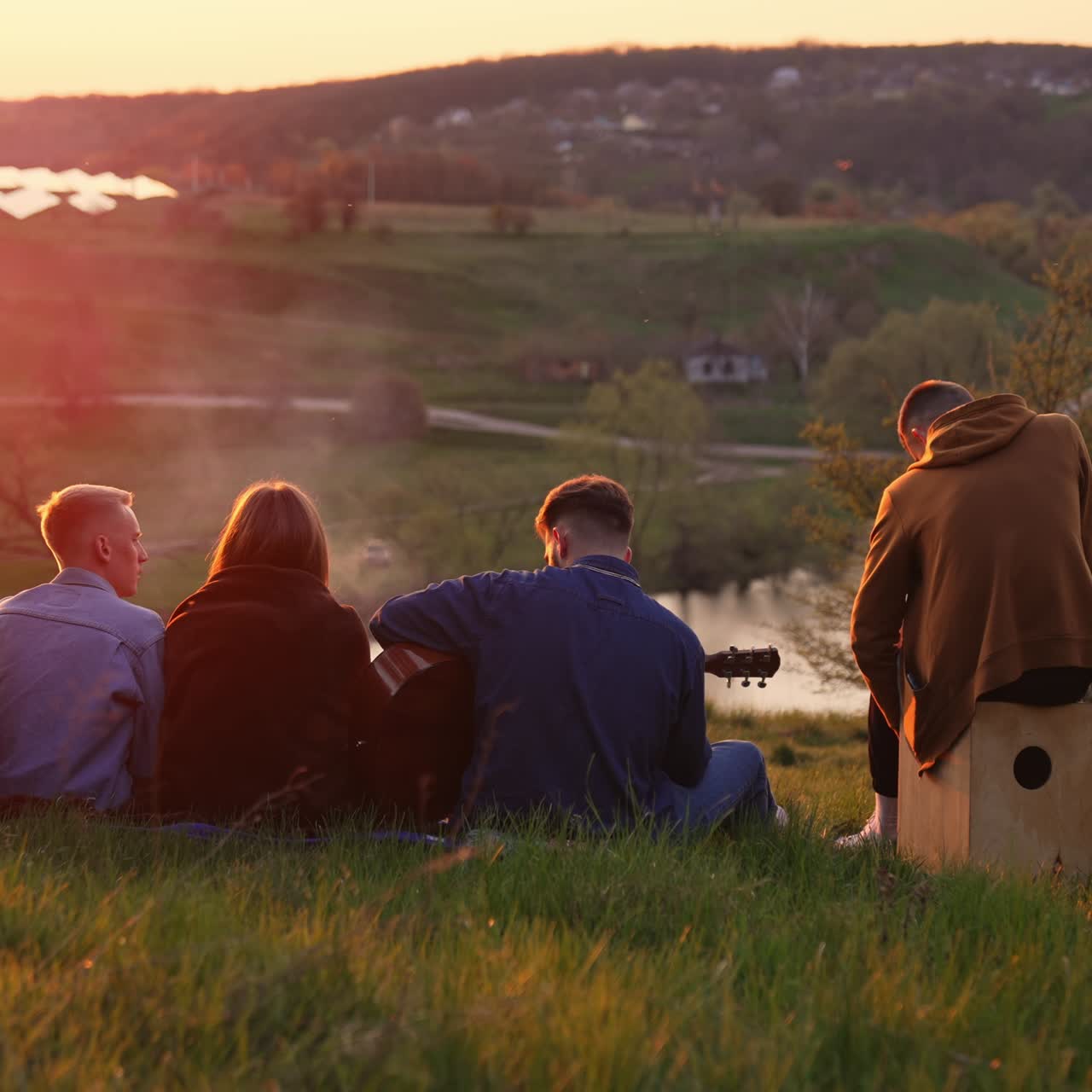 Four young men and woman having good picnic time. Friendly group watching the sunset and singing song along with playing instruments