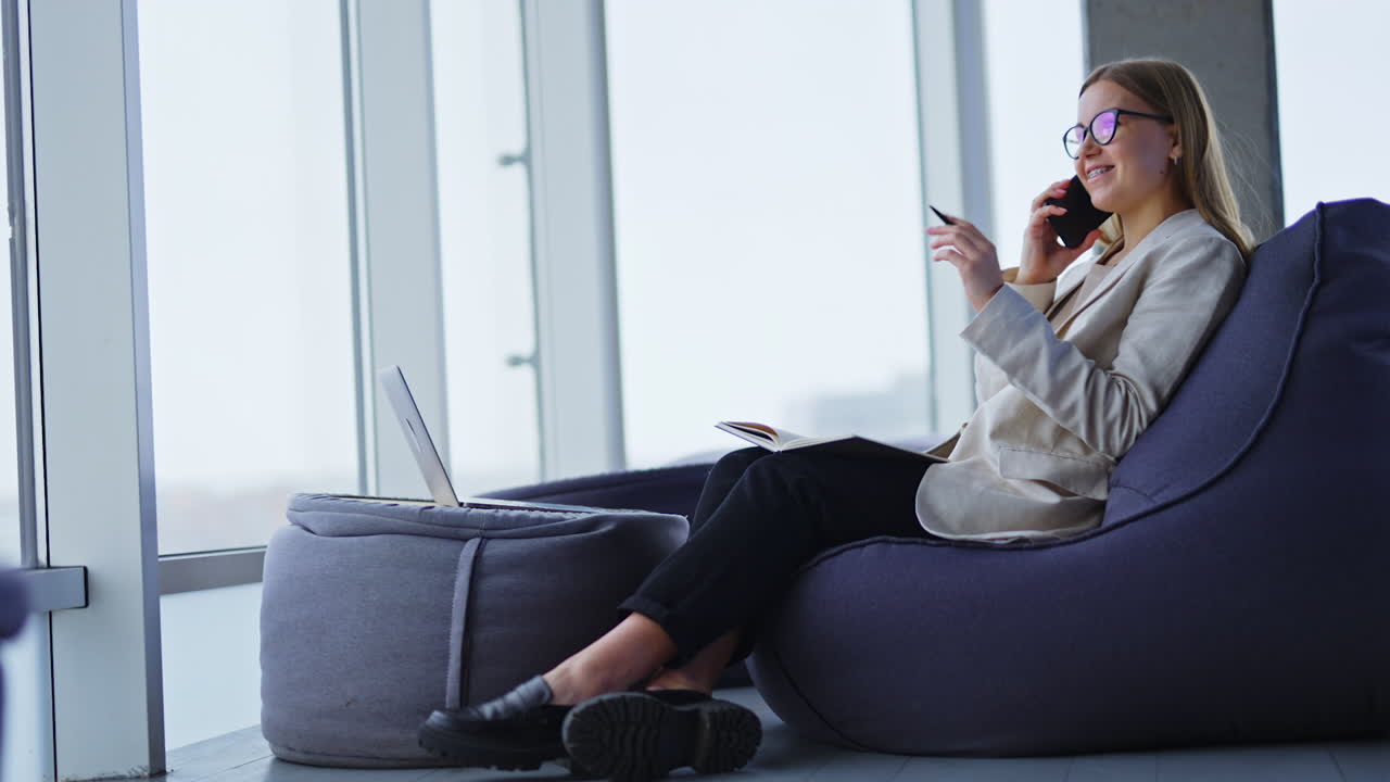 Relaxed businesswoman sitting in a bean bag chair speaks on the phone. Smiling lady has a paper notebook on her knees and pen in her hand.
