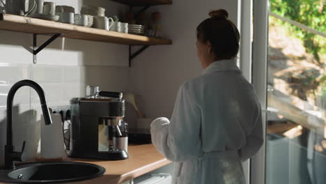 woman makes drink with coffee maker in kitchen. lady in soft bathrobe comes to coffee machine preparing aromatic beverage. concept of relaxed morning