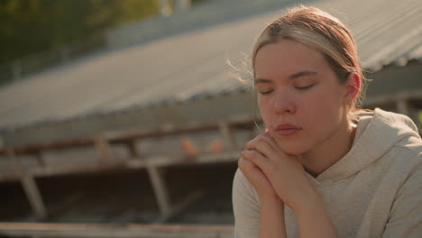 close-up of contemplative woman in casual hoodie seated on rustic stadium bleachers, hands joined near chin, lost in thought, soft lighting highlights her pensive expression