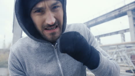 deportista caucásico con capucha gris boxeando a la cámara al aire libre en una fábrica abandonada en una mañana nublada