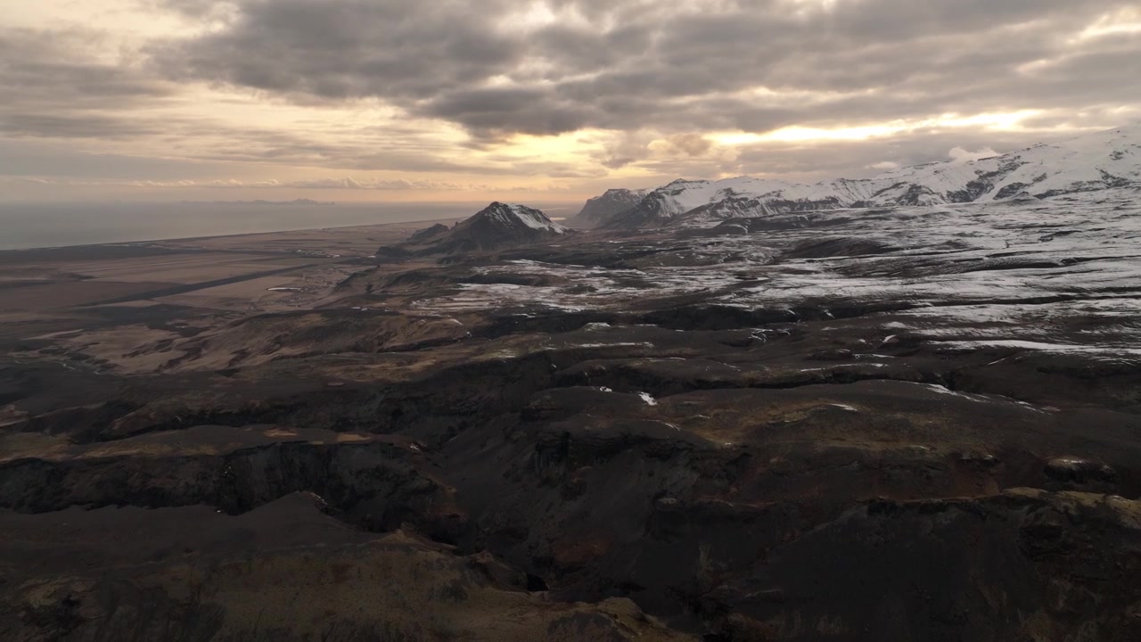 Drone shot of a vast Icelandic landscape at sunset, featuring rugged plains and snow-capped mountains, bathed in warm, dramatic light.
