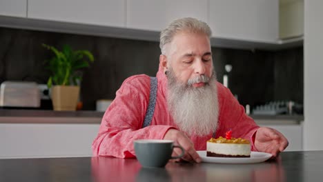 A-sad-elderly-man-with-gray-hair-and-a-lush-beard-in-a-pink-shirt-blows-out-a-candle-on-a-cake-during-his-lonely-birthday-party-in-a-modern-apartment