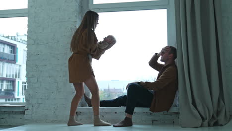 man sits at the windowsill looking at wife waving a baby. tired woman screams at the man holding by the head.