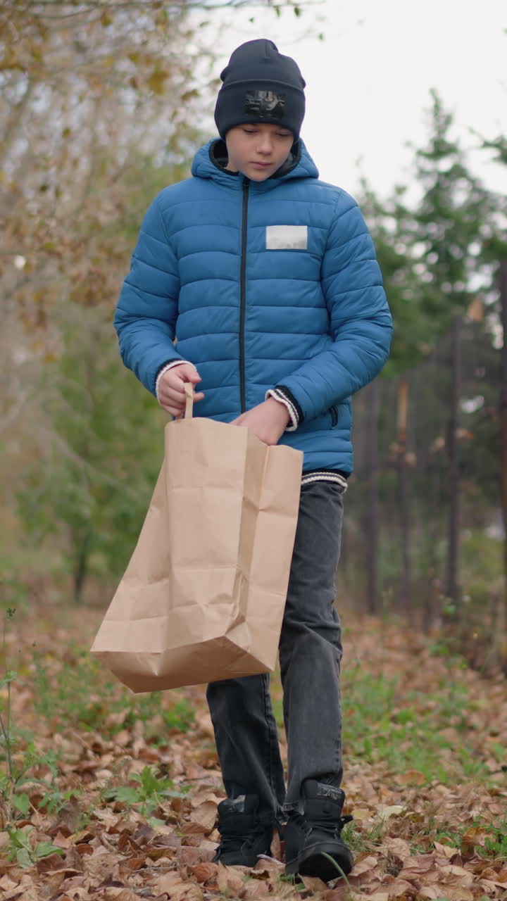 niño en chaqueta azul y gorra se inclina para recoger la hoja oxidada del suelo y la coloca en una bolsa de papel, con la valla de la barra y el edificio en el fondo, rodeado de hojas de otoño