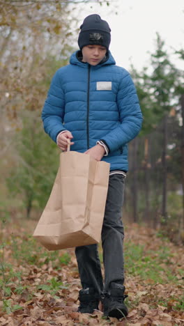 niño en chaqueta azul y gorra se inclina para recoger la hoja oxidada del suelo y la coloca en una bolsa de papel, con la valla de la barra y el edificio en el fondo, rodeado de hojas de otoño