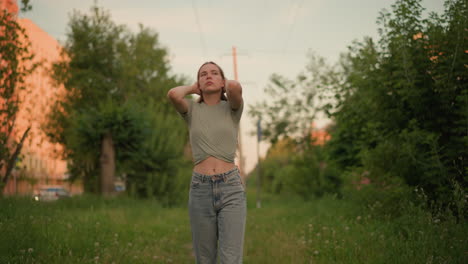 russia lady outdoors with hands placed behind her head, eyes closed in contemplative, moody expression, walking in lush green surroundings and blurred urban background