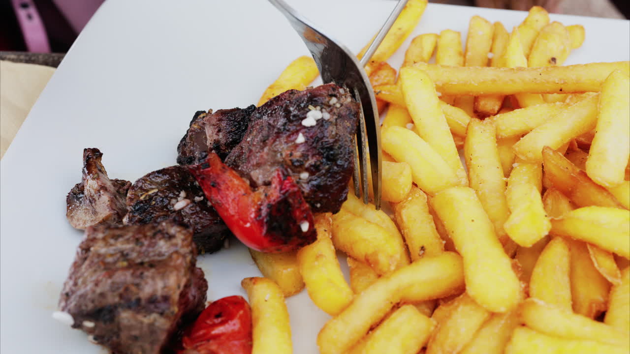 Close up of a woman taking grilled meat off skewers on a white plate with french fries at a restaurant