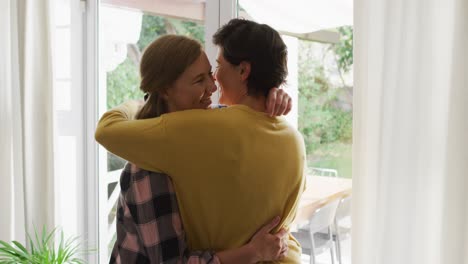 pareja de lesbianas caucásicas abrazándose cerca de la ventana en casa