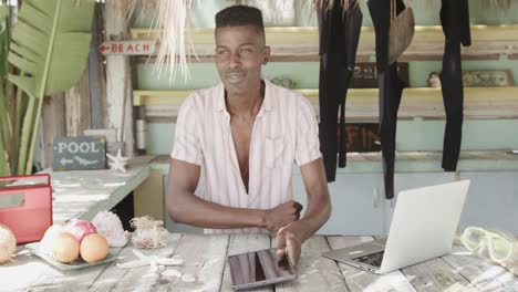 Portrait-of-happy-african-american-man-with-tablet-and-laptop-at-surf-rental-shack,-slow-motion