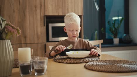 happy little albino boy with white hair color and blue eyes eats his morning breakfast while sitting at the table in a modern apartment kitchen. happy boy eats breakfast from a plate using a spoon while sitting on a white chair at the table