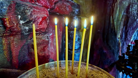 thin candles burn brightly in a sand-filled holder, set against a vivid, textured wall of red, yellow, and blue tones - pan right shot