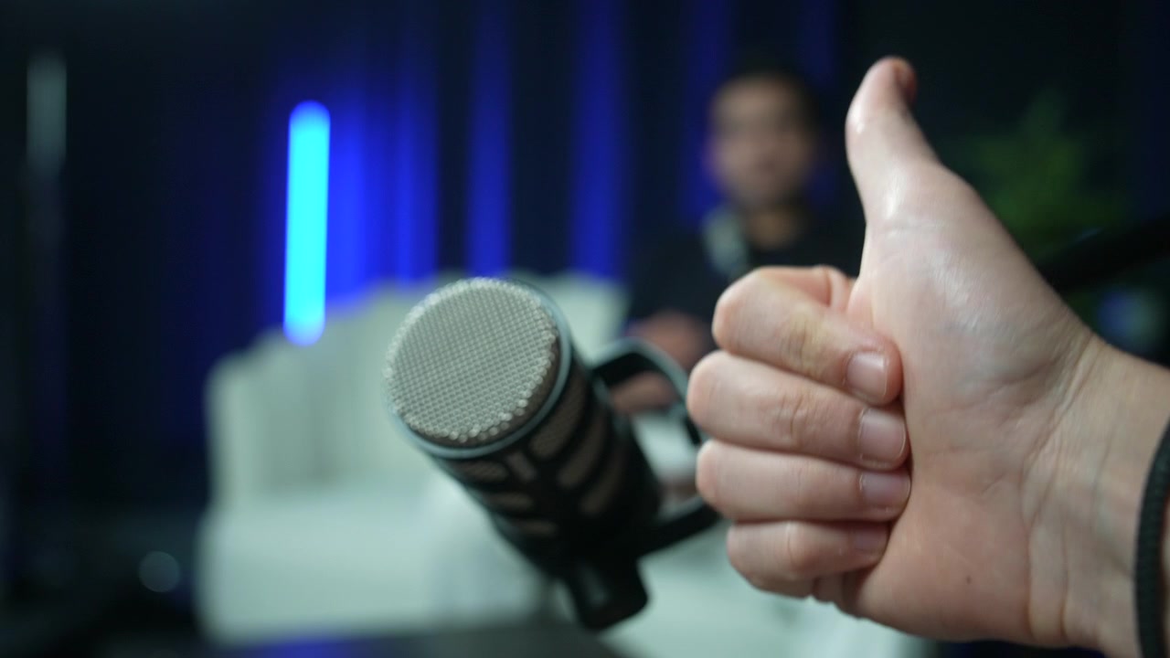Mão faz sinal de positivo perto do microfone em um ambiente de entrevista de estúdio descontraído, ajustando o suporte do microfone