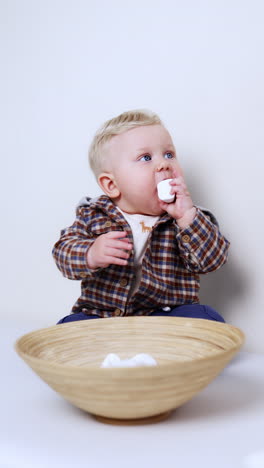 cute little blond baby boy sits at a big bowl. kid takes marshmallows from the dish and puts to his mouth. white backdrop. vertical video.