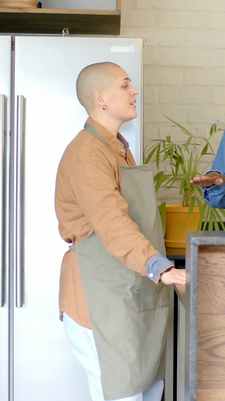 Vertical video: Talking woman in olive apron responding to coworker gesturing at kitchen counter