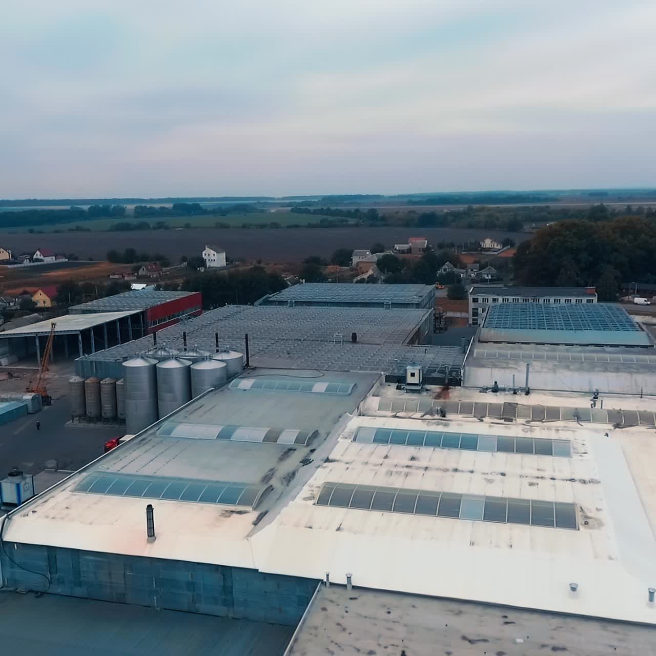 Industrial premises with metal constructions and windows on top. Countryside area with private houses, roads and fields. Top view