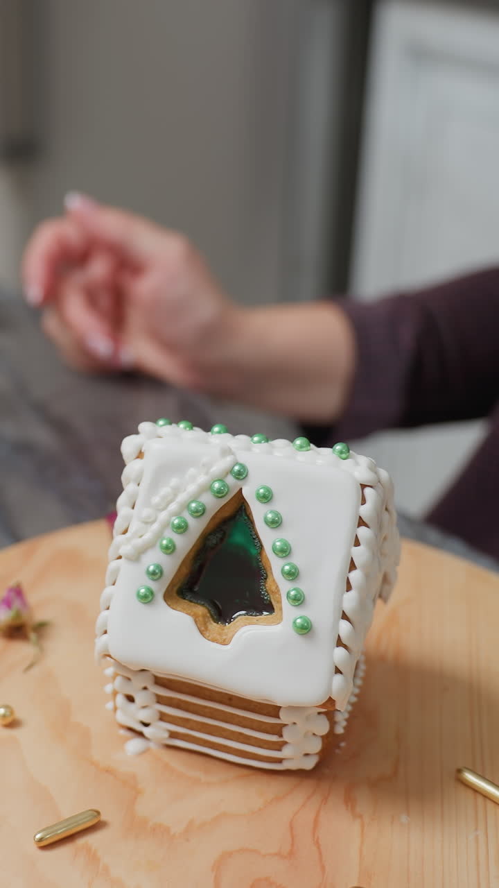 primer plano de una casa de pan de jengibre decorada con hielo blanco y cuentas verdes en una bandeja de madera, pequeñas decoraciones doradas, flores secas, un frasco de cuentas verdes y una planta en maceta