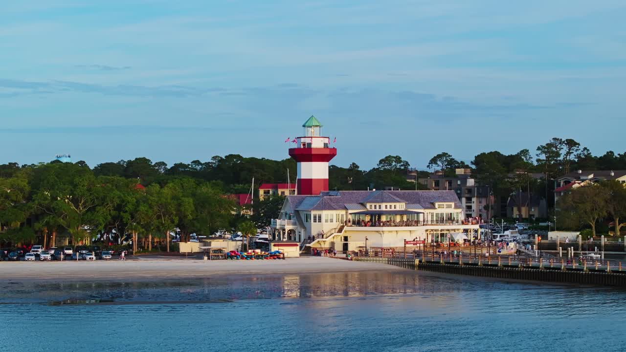 High drone ascend over turquoise harbor with boats docked around town pier, red and white light house, Harbour Town Pier South Carolina USA