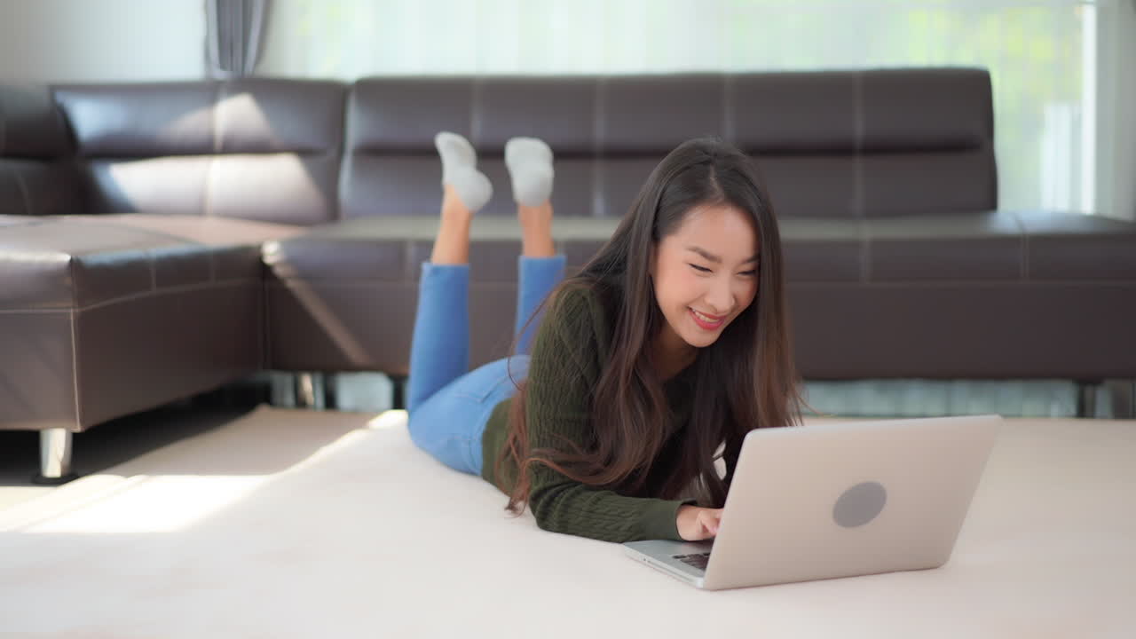 Asian model laying on floor with laptop computer. Woman smiling as she uses Internet to communicate with friends. Happy attractive lady relaxing at home in living room. Self isolation