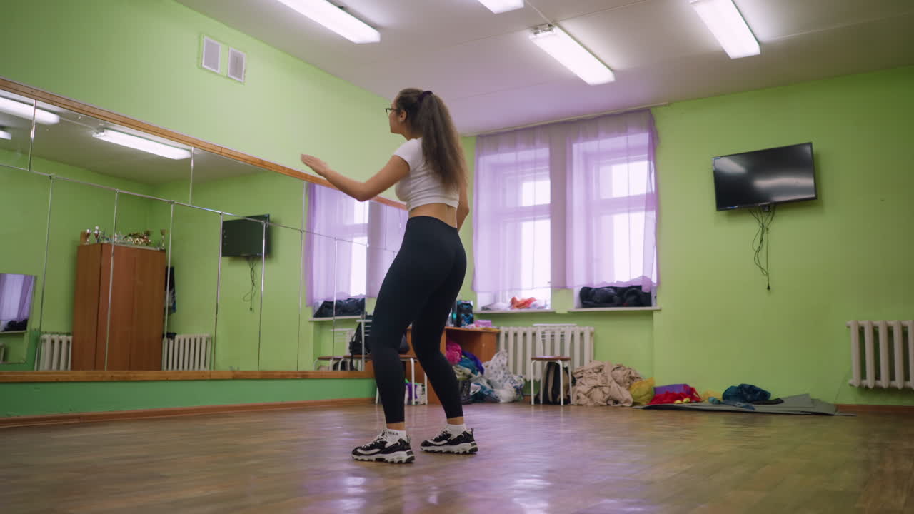 Girl in white top and black leggings dancing in green painted room with large mirror wall, wooden floor, and scattered items, showing graceful movement and energetic posture during indoor practice