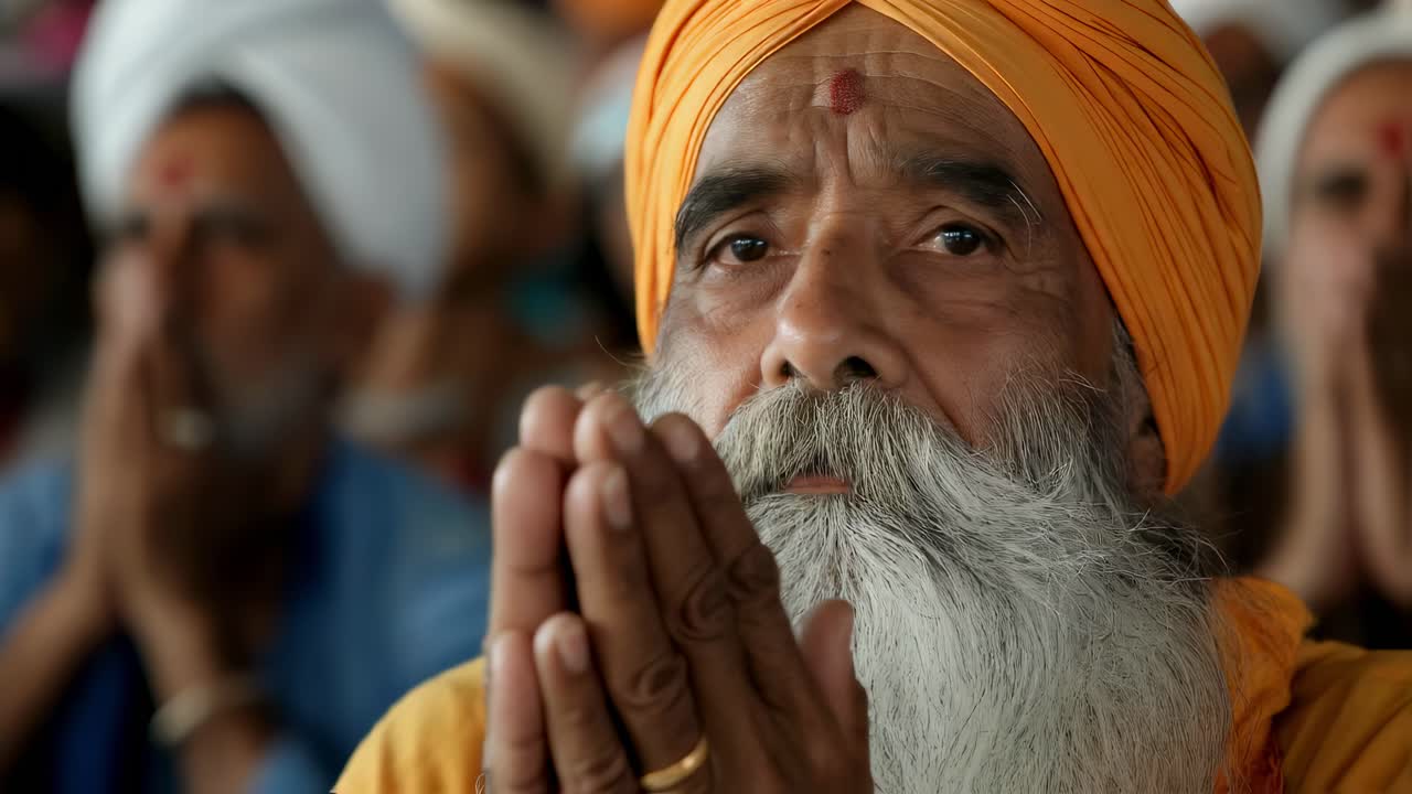 Bearded sikh religious leader wearing orange turban is praying with closed eyes and joined hands, surrounded by blurred followers in Amritsar Golden Temple