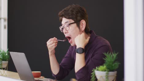 Asiatischer-Mann,-Der-Einen-Snack-Isst-Und-Dabei-Auf-Den-Laptop-Schaut,-Während-Er-In-Einem-Café-Sitzt