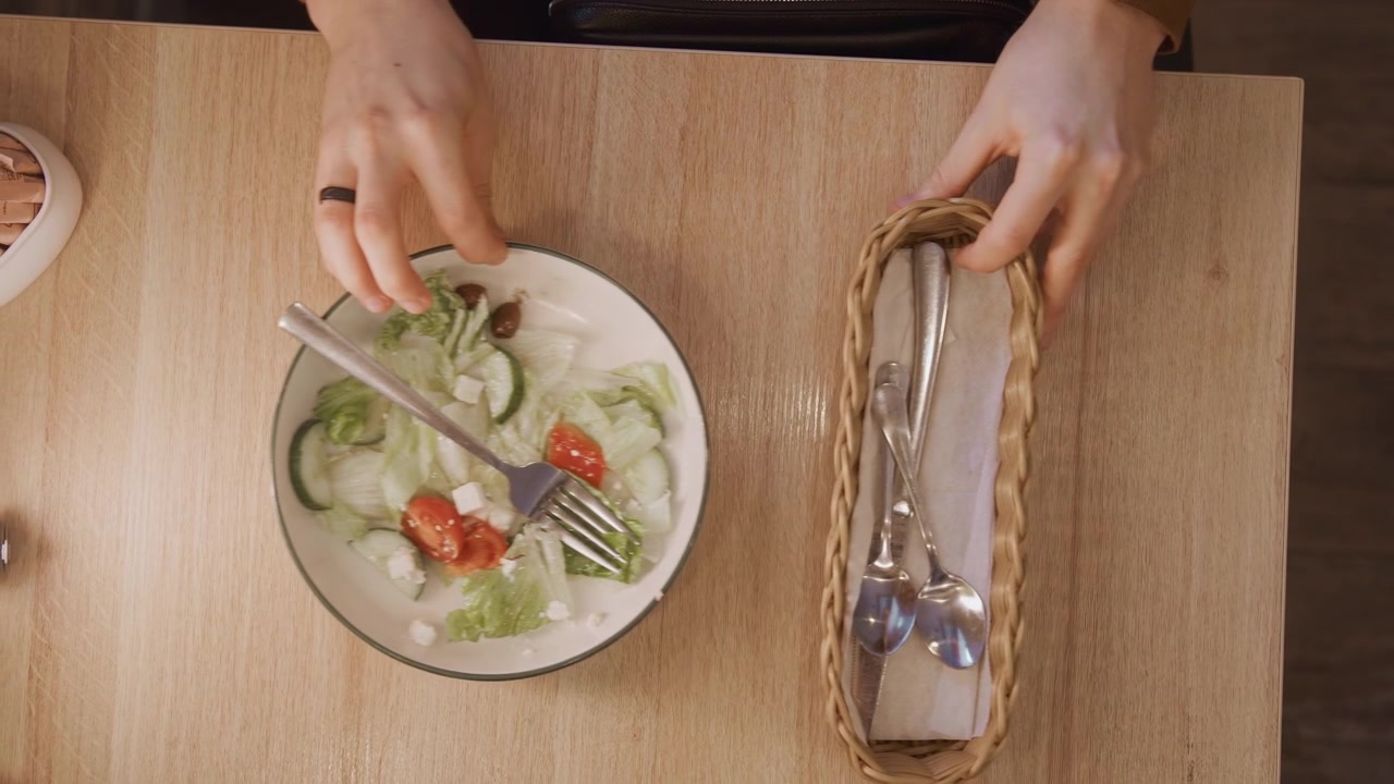 Woman hands interact with sketchbook and utensils on wooden table. salad bowl pencil cutlery basket are arranged as woman moves objects around. scene is overhead view of workspace items