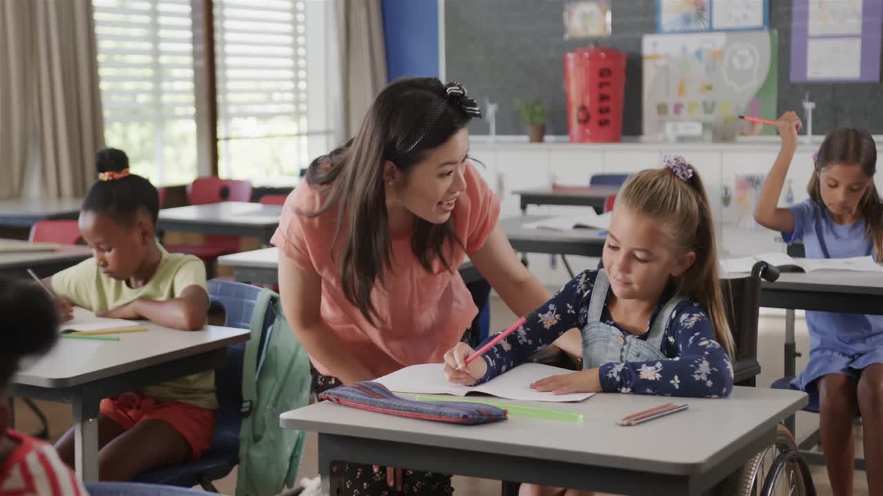 una feliz maestra diversa hablando con una niña en silla de ruedas en una clase de escuela primaria, cámara lenta