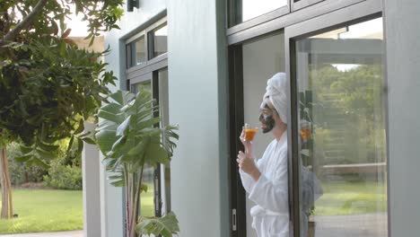 happy caucasian woman with mask drinking tea at window and looking at garden in slow motion