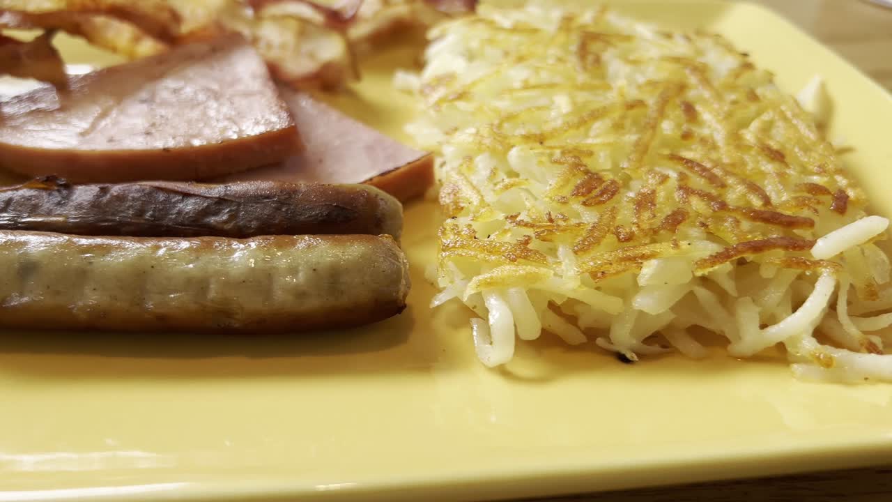 Two greasy sausages, savory slices of ham, golden hash browns, and crispy bacon arranged on a vibrant yellow plate, creating a hearty and indulgent American breakfast feast panning slow motion shot