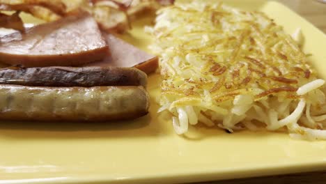 two greasy sausages, savory slices of ham, golden hash browns, and crispy bacon arranged on a vibrant yellow plate, creating a hearty and indulgent american breakfast feast panning slow motion shot