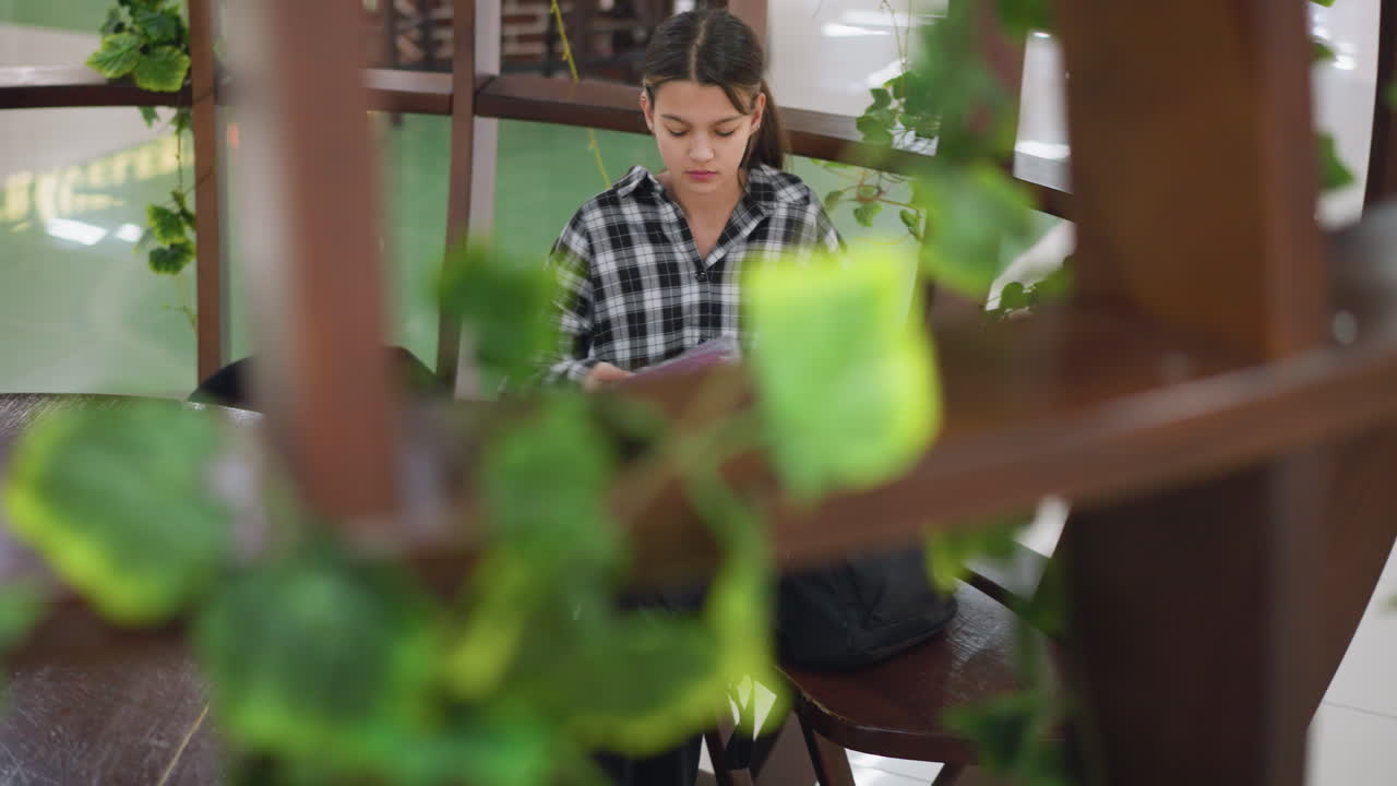 joven se sienta en un asiento de madera en un área decorada, mirando estresado mientras revisa su bolsa de la escuela negra, la vegetación rodea el espacio acogedor, creando una atmósfera reflexiva