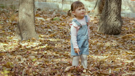 young asian girl points at autumn leaves, smiles, before playfully kicking them