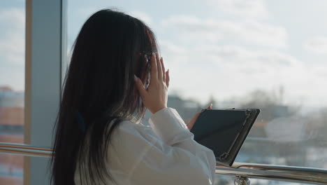 Rear view of woman in white shirt adjusting glasses while operating tablet near glass railing, with outdoor scene softly blurred in background and sunlight filtering through large windows