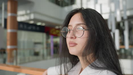 young woman with long dark hair and eyeglasses looking thoughtful while standing inside modern indoor space with soft daylight and blurred commercial background