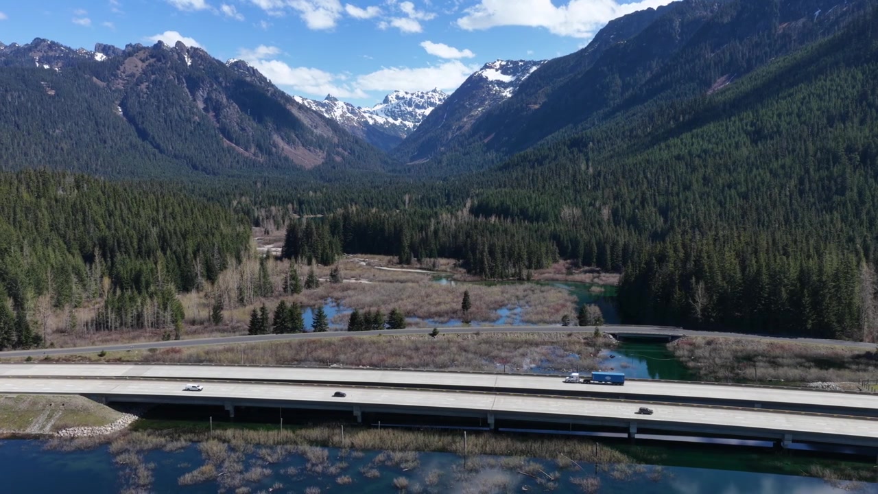 Ponte autostradale che attraversa il fiume in un'ampia valle di montagna, cime delle Cascate innevate che si ergono sopra una fitta foresta sempreverde con auto in viaggio sotto un cielo azzurro vivido. Washington, USA. Ampia ripresa aerea