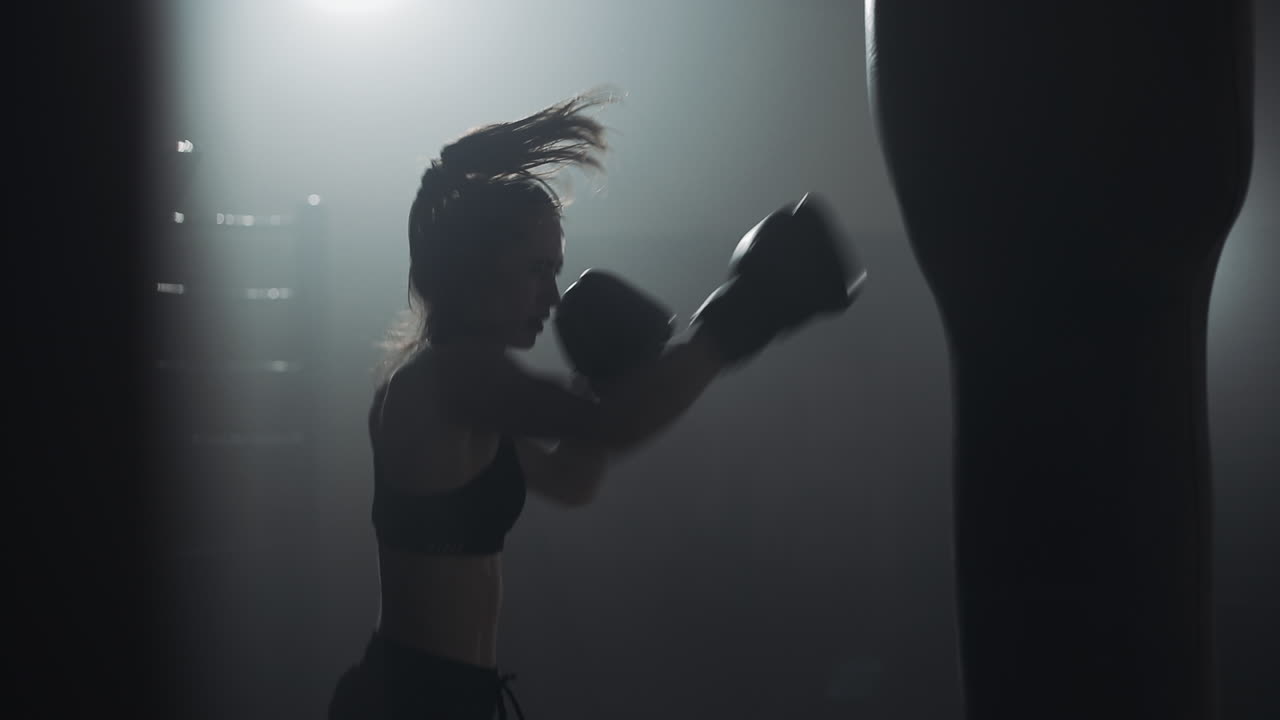Pugile donna che si allena con il sacco da boxe in un'illuminazione drammatica