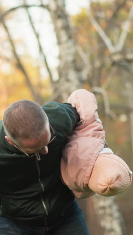un momento alegre capturado en un parque al aire libre soleado donde un hombre con una chaqueta verde gira juguetónamente a una niña con una jaqueta rosa y pantalones vaqueros azules, ambos sonriendo y disfrutando de la atmósfera alegre