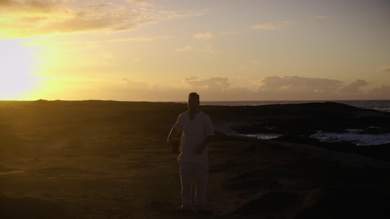 A man strides forward along a rugged coastal path, backlit by the golden sunrise as waves crash in the distance and the horizon glows warmly