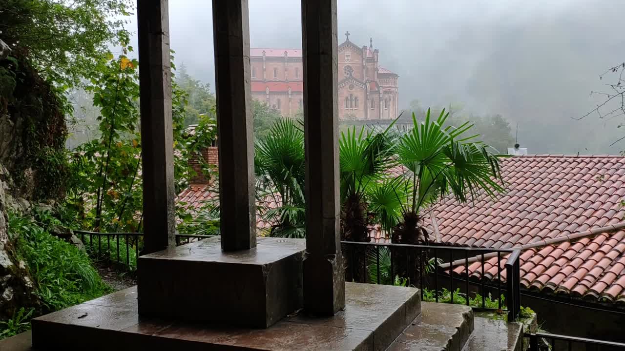 Rainy stone monument near Basilica de Covadonga with palm trees and rooftop view