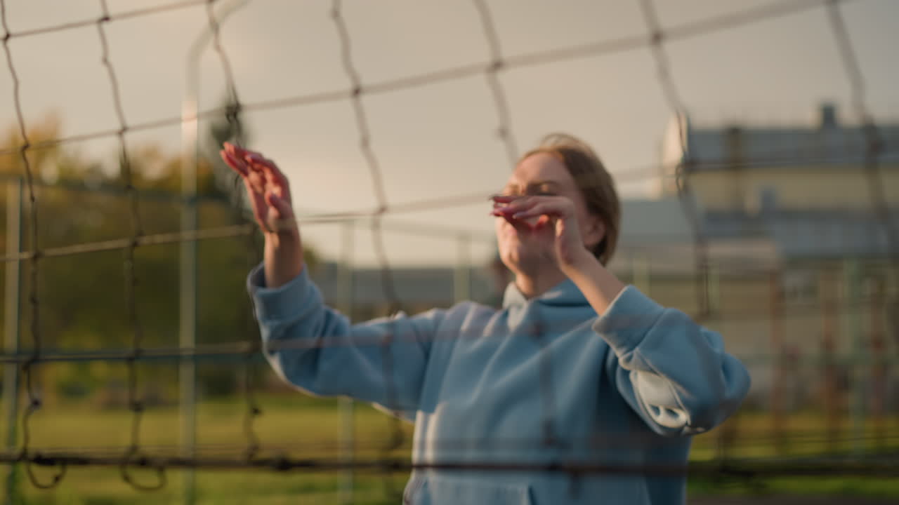 primer plano de la red de voleibol con el jugador visible en el fondo preparándose para un servicio, enfoque suave en la red, mientras que la vegetación y el edificio aparecen borrosos