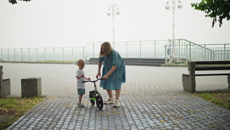 a mother bends down to assist her child by holding his bicycle as he climbs it, with a blur view of a little boy behind close to an iron rail