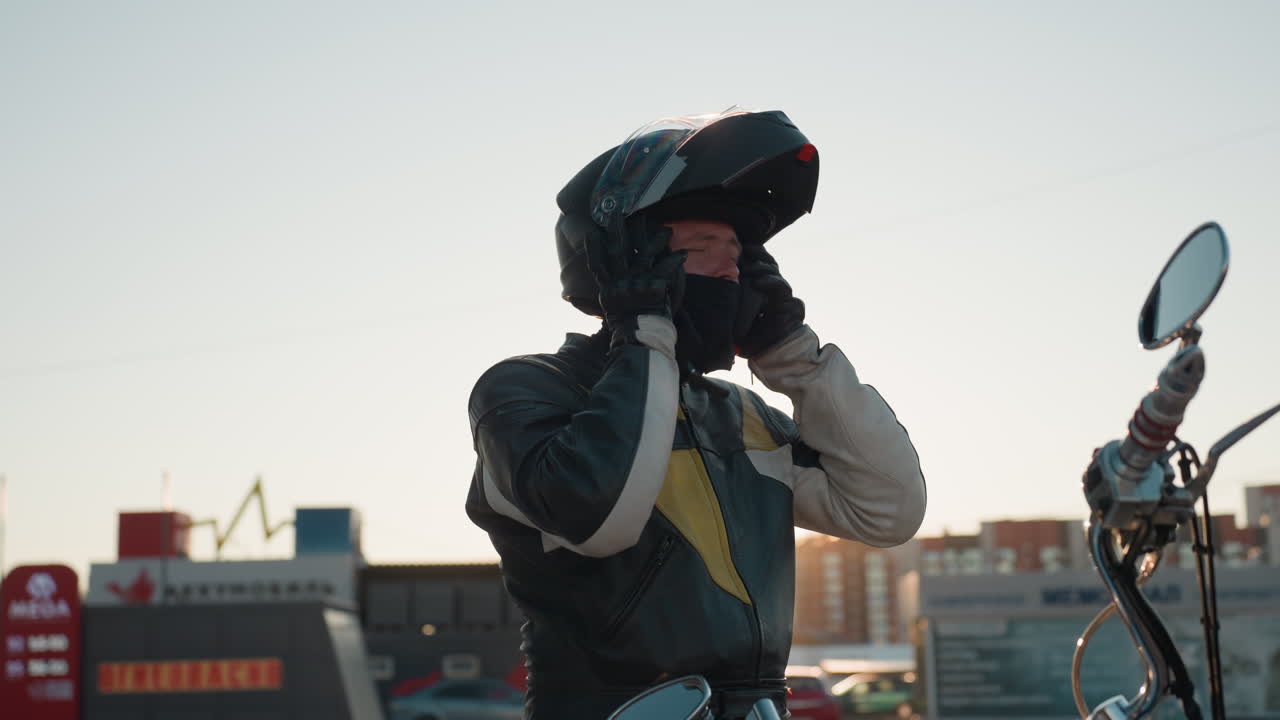 Rider unbuckles helmet preparing to remove it under bright sunlight wearing protective leather jacket standing near motorcycle with urban background