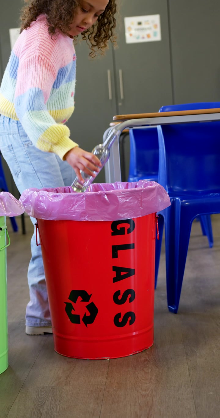 Vertical video: African American girl in pastel top with bottles walking to GLASS bin for recycling
