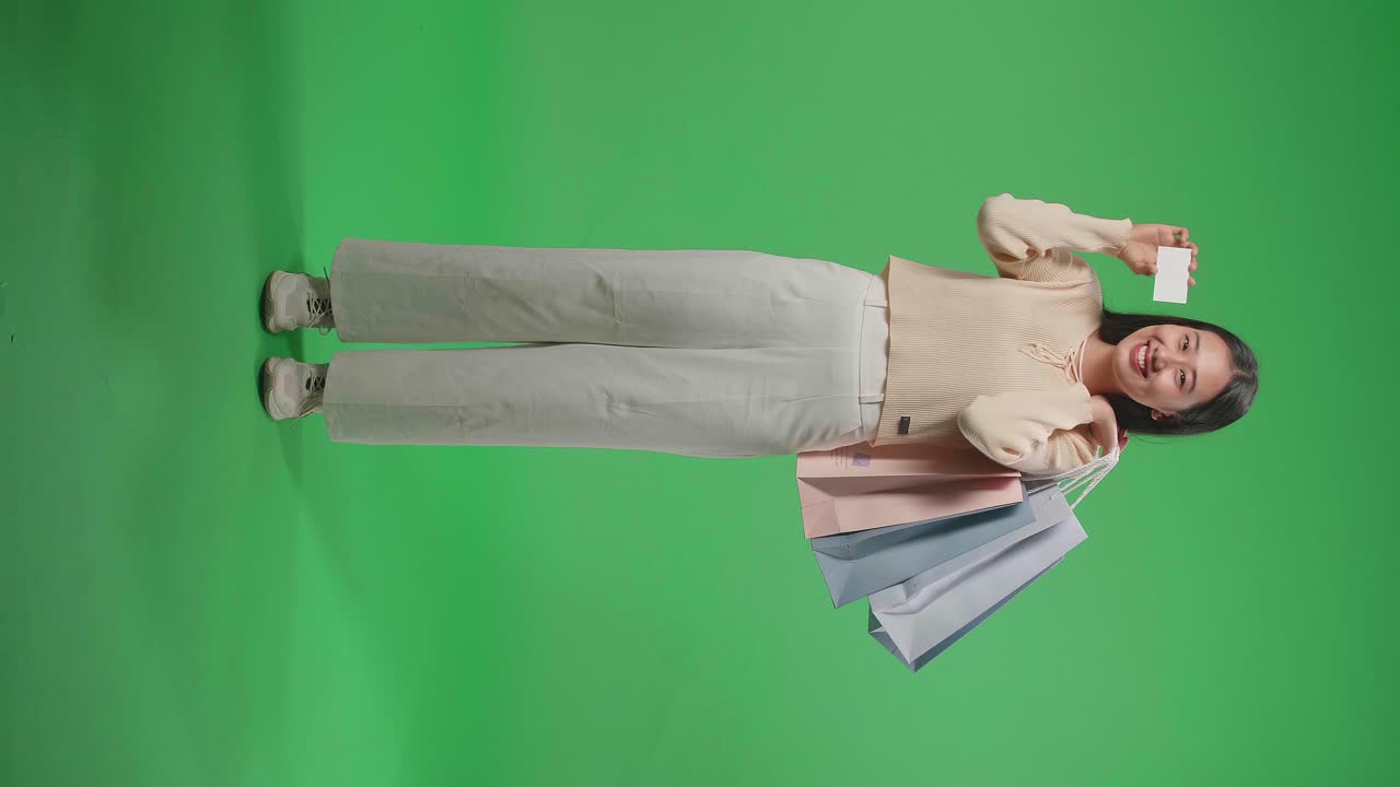 Full View Of An Asian Shopping Woman Holding A White Card And Shopping Bags Up Before Smiling To Camera While Standing In Front Of Green Screen